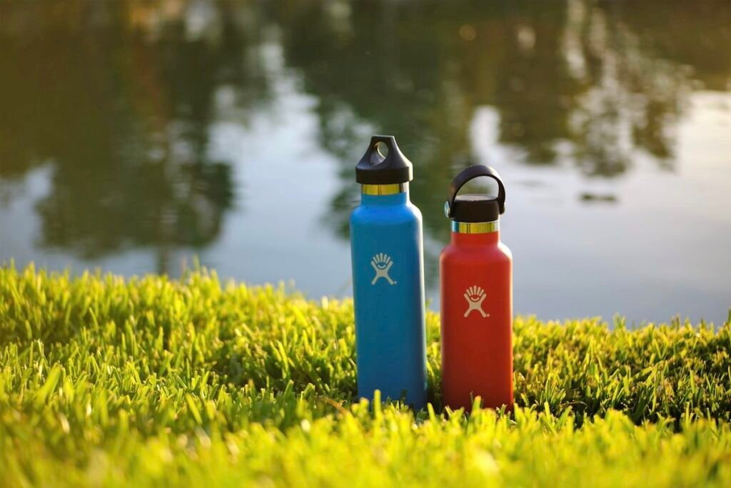 Twee geïsoleerde RVS drinkflessen in blauw en rood buiten bij een meer, die lang koud of warm houden
